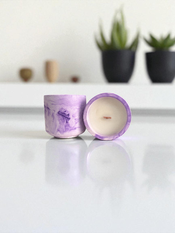 Two purple marbled candles on a white surface with plants in the background