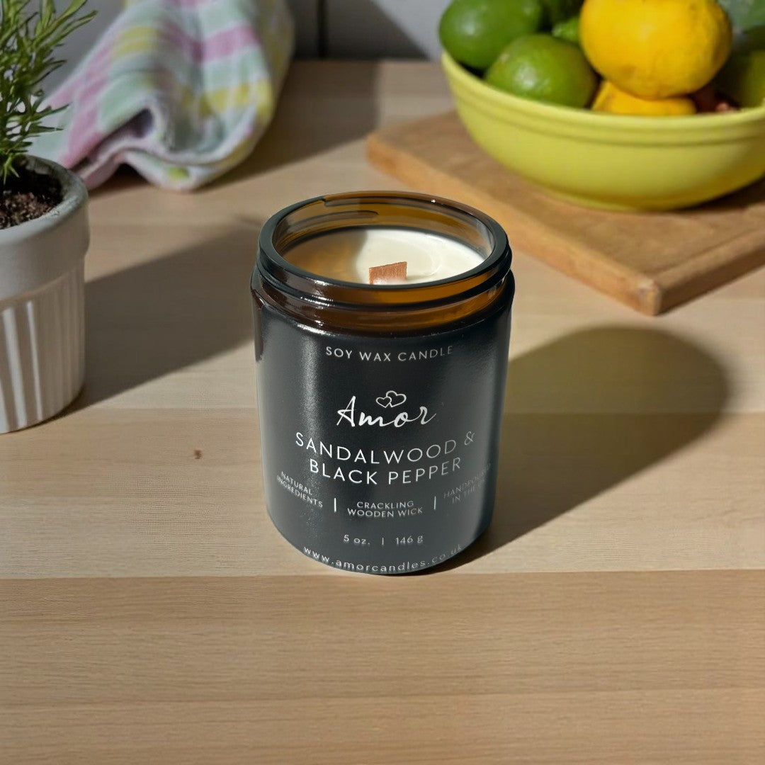 Amor Sandalwood & Black Pepper candle on a wooden surface with a bowl of fruit and plant in the background