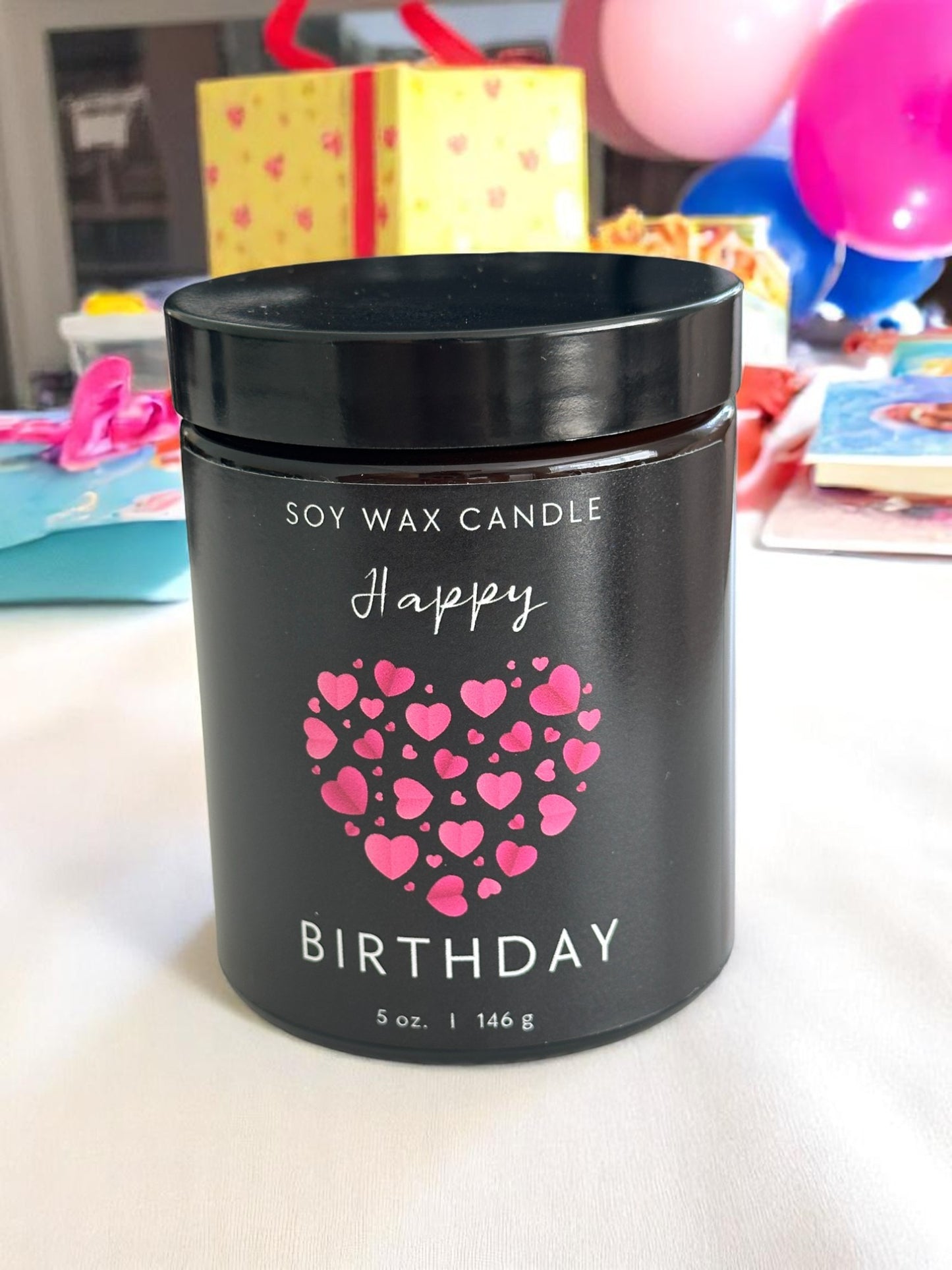 Black soy wax candle with pink hearts and 'Happy Birthday' text on a blurred background of balloons and gifts.
