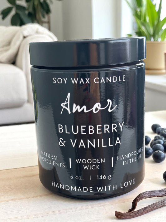 Black soy wax candle labeled 'Amor Blueberry & Vanilla' on a wooden surface with vanilla beans and blueberries.